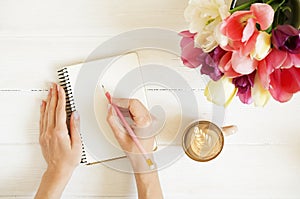 Overhead shot of woman hands drawing, writing with pencil in open notebook, drinking coffee on white wooden table. Beautiful tulip