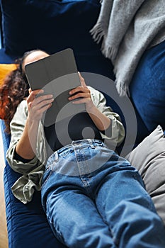 Overhead Shot Of Young Woman Relaxing At Home Lying On Sofa Reading Book On Digital Tablet