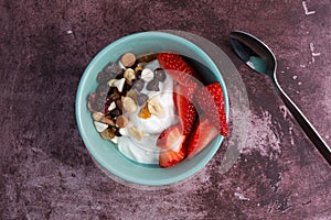 Overhead view of a bowl with plain Greek yogurt and a candy trail mix and strawberries on a marron tabletop with a spoon to the