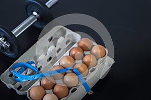 Overhead view of brown chicken eggs in an open egg carton, dumbbell with weighing scale and measuring tape on black table