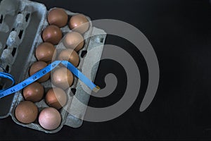 Overhead view of brown chicken eggs in an open egg carton, dumbbell with weighing scale and measuring tape on black table