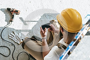 overhead view of builder in protective helmet using smartphone with blank screen at construction