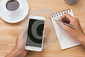 Overhead view of businessman hand holding smart phone while writing on notepad by camera and coffee on table