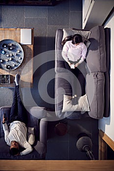 Overhead View Of Couple Relaxing On Lounge Sofa At Home Watching TV And Using Mobile Phone