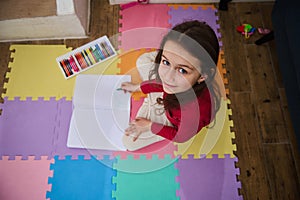 Overhead view of a cute little child girl smiling looking at camera, drawing with pastel pencils, sitting on a colorful