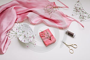 Overhead view of essential beauty items, top view of pink silk fabric, natural soap, flowers and decorative scissors