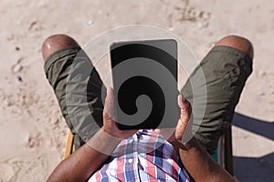 Overhead view of man holding digital tablet while sitting on deck chair at the beach