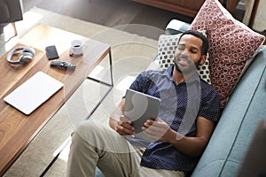 Overhead View Of Man Lying On Sofa At Home Watching Movie On Digital Tablet