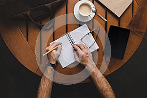 overhead view of man writing in notepad at wooden table with digital tablet, coffee