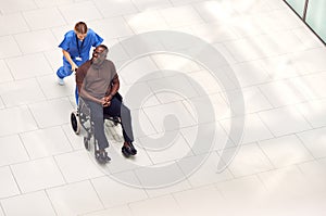 Overhead View Of Nurse Wheeling Male Patient In Wheelchair Through Lobby Of Modern Hospital Building