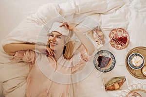 Overhead view of radiant young caucasian girl lying in snow-white bed among plates with food.