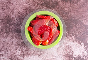 Overhead view of a small green bowl filled with slices of fresh strawberries on a dark red mottled tabletop illuminated with