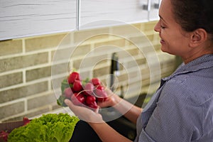 Overhead view woman holding fresh radish, standing by kitchen counter with a cardboard box full of delivered vegetables