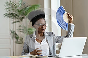 Overheated african businesswoman wave fan drink water sitting at office desk with laptop or at home