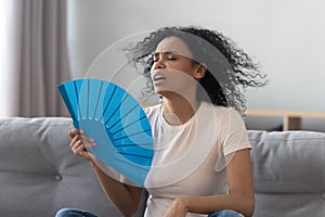 Overheated african young woman feeling hot waving fan at home