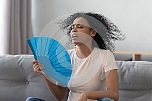 Overheated african young woman feeling hot waving fan at home photo