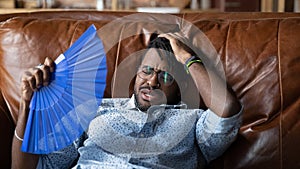 Overheated afro american man recline on couch use hand fan photo