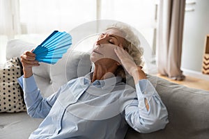 Overheated elderly female leaned on couch refreshing herself with fan