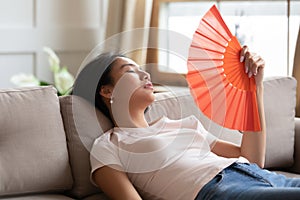 Overheated exhausted Asian woman waving paper fan, sitting on couch