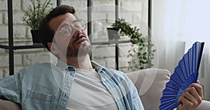 Overheated man sitting on sofa hold fan cooling himself