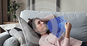 Overheated or sick young woman waving fan lying on sofa