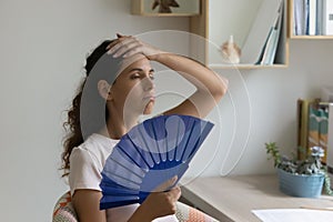 Overheated woman sit indoors feels overheated holding handheld blue fan