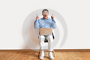 Overjoyed asian man using pc and pointing fingers up, sitting on chair against white studio background wall, free space