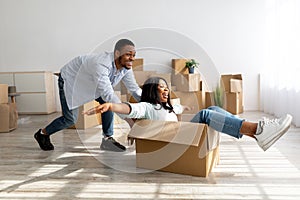 Overjoyed black spouses having fun while moving to new home, celebrating relocation, riding in cardboard box