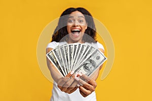 Overjoyed black woman holding money cash in hands, standing over yellow studio background, selective focus