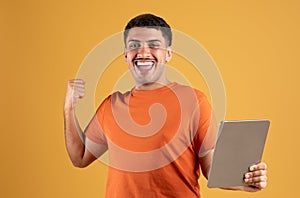 Overjoyed brazilian man holding digital tablet and making victory sign, celebrating success on yellow studio background