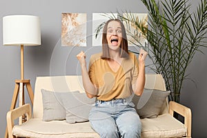 Overjoyed brown haired woman wearing beige T-shirt and jeans sitting on sofa in living room hearing positive mood clenched fists