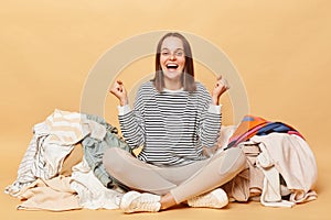 Overjoyed Caucasian woman posing near heap of multicolored unsorted clothes isolated over beige background clenched fists