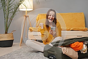 Overjoyed Caucasian young adult woman packing suitcase at home using mobile phone for buying tour or tickets for having journey