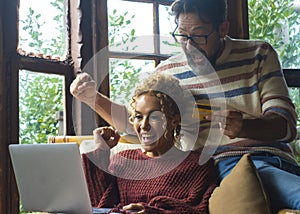 Overjoyed couple exult together in front of laptop sitting at home on the sofa. Excited people man and woman celebrate holding