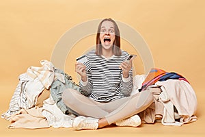 Overjoyed crazy Caucasian woman posing near heap of multicolored unsorted clothes isolated over beige background holding mobile