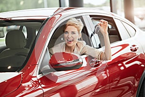 Overjoyed driver woman smiling and showing new key while sitting in car showroom