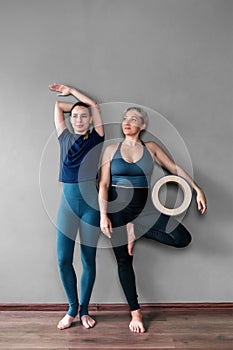 Overjoyed full-length female yoga trainers in sportswear stand barefoot near the wall, one holds a wooden circle in her hand