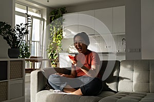 Overjoyed happy Asian woman screaming sitting on sofa with phone celebrating sports team victory