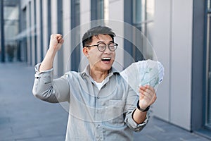 Overjoyed korean man holding lots of money and gesturing YES, asian male with fan of euros, standing outdoors