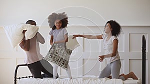 Overjoyed little african ethnicity child girl fighting pillows with parents.
