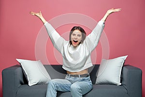 Overjoyed lucky young girl celebrating win with raised arms, screaming with euphoria, sitting on sofa