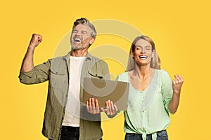 Overjoyed middle aged couple shaking fists holding laptop computer and gesturing yes, celebrating great news