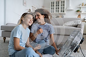 Overjoyed mother and teen daughter enjoying fresh cold air from electrical fan, sitting on floor