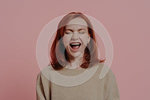 Overjoyed red-haired woman laughing positively, with closed eyes, isolated on pink background photo