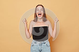 Overjoyed surprised brown haired young woman wearing black top and jeans standing isolated over beige background celebrfating her