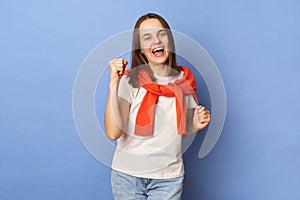 Overjoyed woman wearing white t-shirt and jumper over neck standing isolated over blue background clenched fists celebrating her