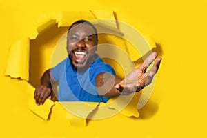 Overjoyed young black man stretching open palm through hole in torn yellow paper background, taking or giving something