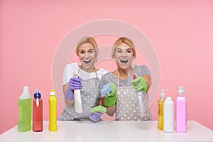 Overjoyed young blonde housewives in basic t-shirts and aprons keeping bottles with household chemicals and looking cheerfully at photo