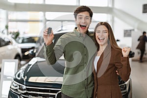 Overjoyed young Caucasian couple making YES gesture, showing car key, buying new auto at dealership, copy space