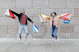 Overjoyed young diverse couple with gift bags jumping near grey brick wall, going shopping together